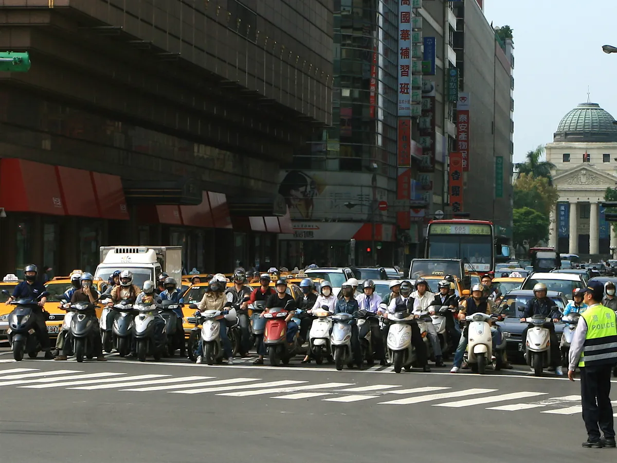 台北館前路口,一整排機車騎士戴著安全帽停在斑馬線前等紅燈,旁邊有交通警察指揮
